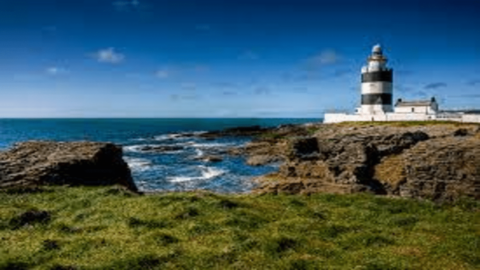 A black and white lighthouse stands near the rocky coast, surrounded by grass, with the ocean and blue sky in the background.