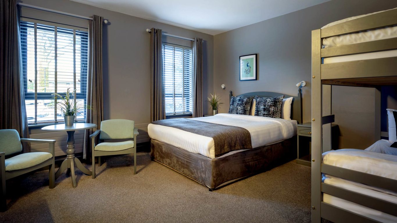 A hotel room with a double bed, bunk beds, two chairs with a small round table by the window, gray walls, and framed artwork above the bed.