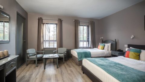 A hotel room with two single beds, a seating area with two chairs and a table, a vanity, and two windows with blinds and curtains.