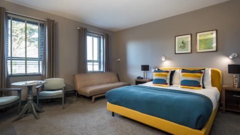 A modern hotel room with a yellow bed, blue bedding, a beige sofa, two windows, a small round table with chairs, and framed artwork on the wall.