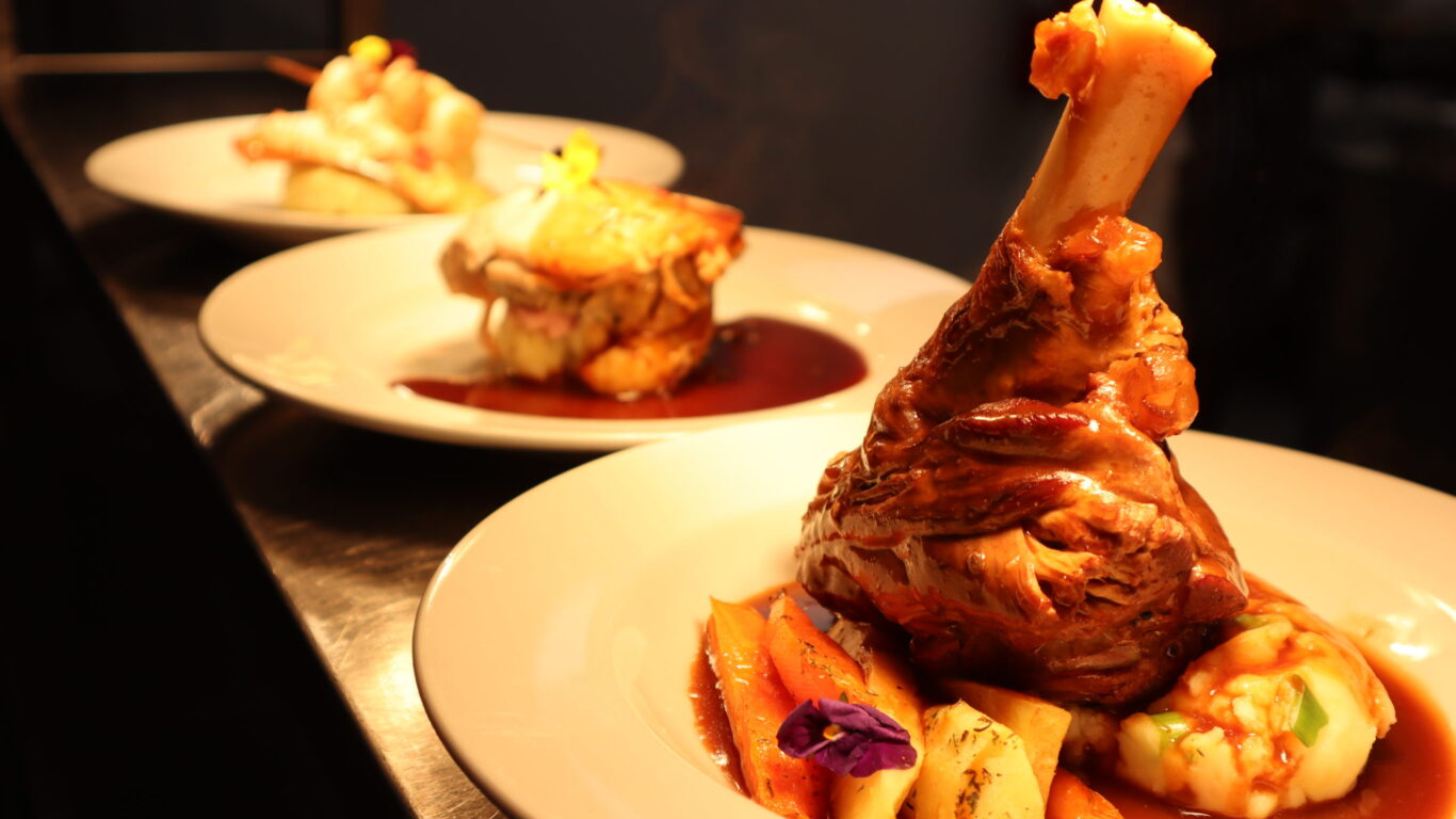 A plated lamb shank with vegetables and sauce in the foreground, with two similar plated dishes in the background on a counter.