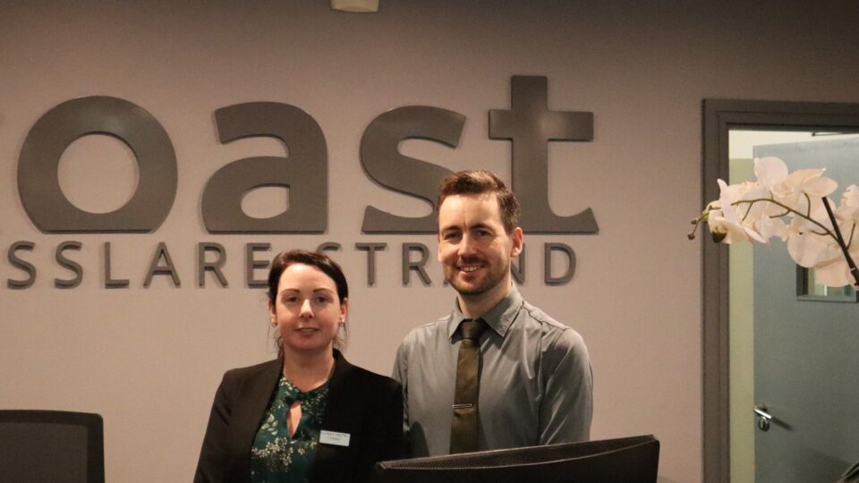Two hotel reception staff stand behind a front desk with a computer monitor, beneath a sign that reads 