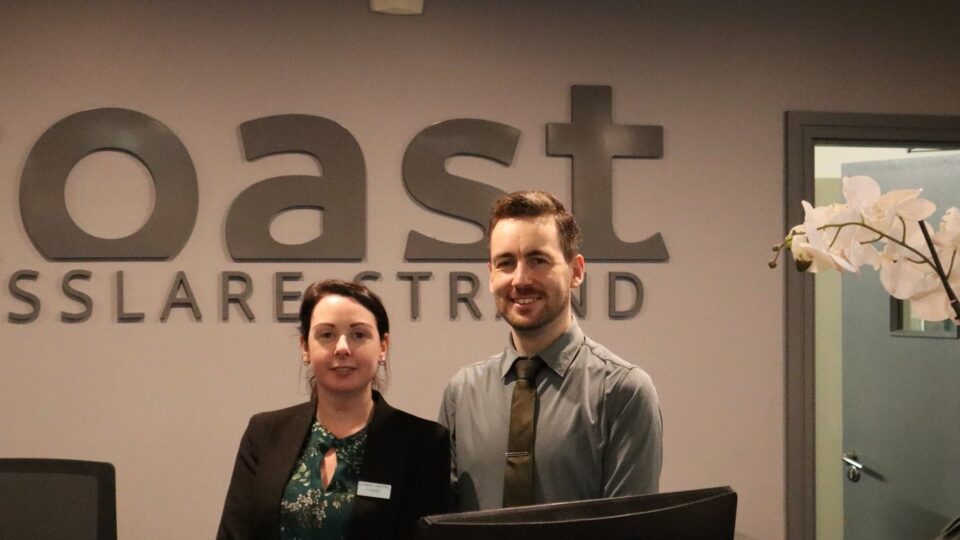 Two hotel reception staff stand behind a front desk with a computer monitor, beneath a sign that reads 