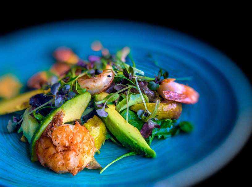 Shrimp, sliced avocado, greens, and microgreens arranged on a blue plate against a black background.