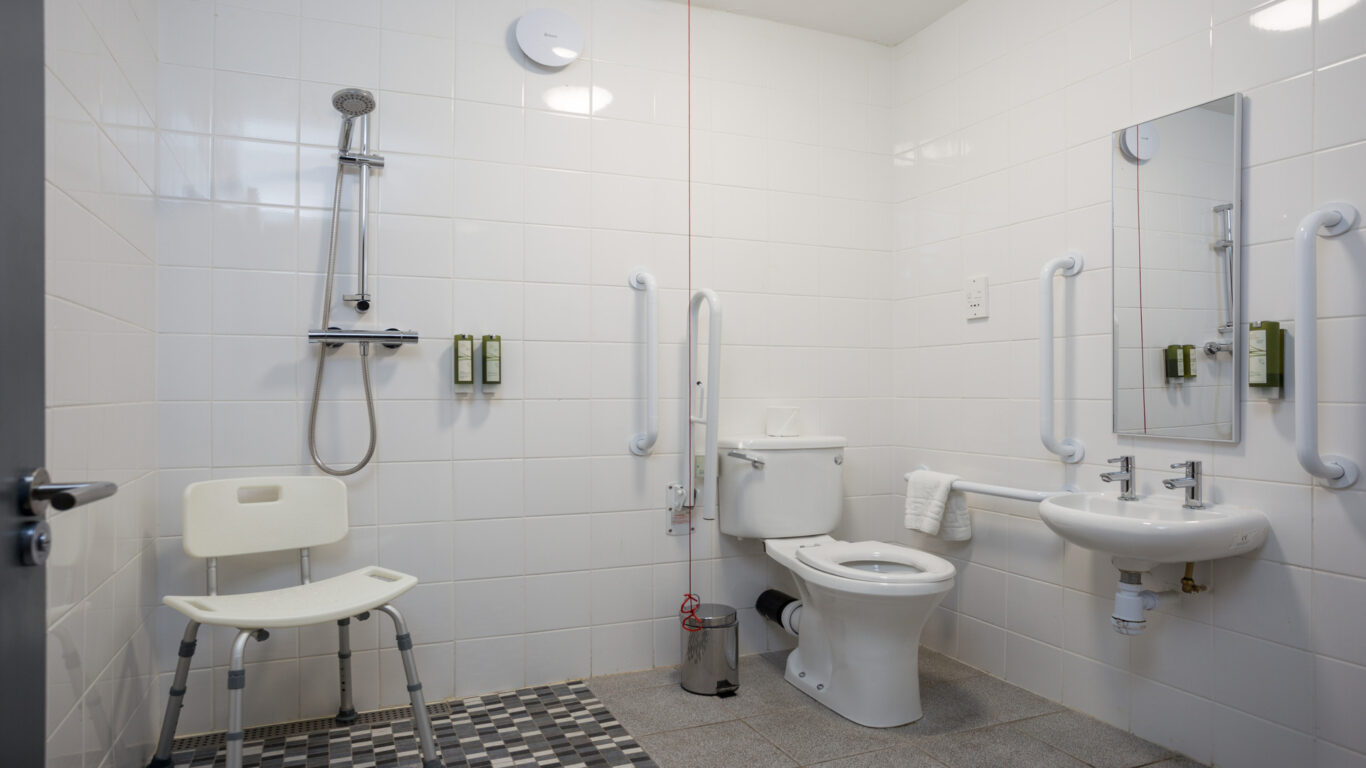 Accessible bathroom with a shower, shower chair, toilet, grab bars, sink, mirror, and amenities mounted on white tiled walls.
