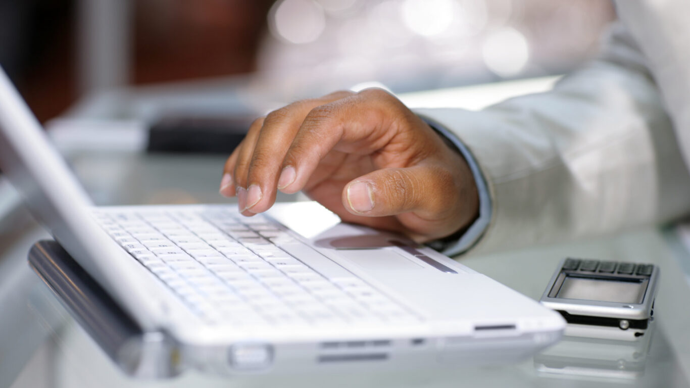 A person&rsquo;s hand types on a white laptop keyboard with a smartphone placed beside the laptop on a glass table.