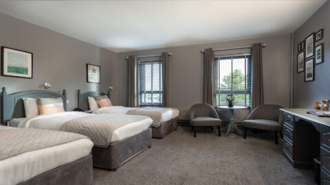 A hotel room with three single beds, two armchairs around a small table, framed art on gray walls, and windows with gray curtains and blinds.