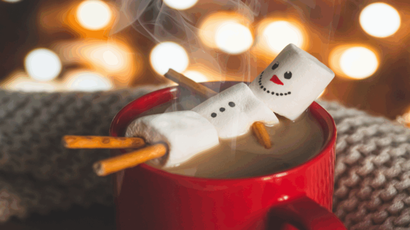 A red mug holds a hot drink topped with marshmallows arranged like a snowman, with pretzel sticks for arms and legs, in front of a cozy, blurred background.