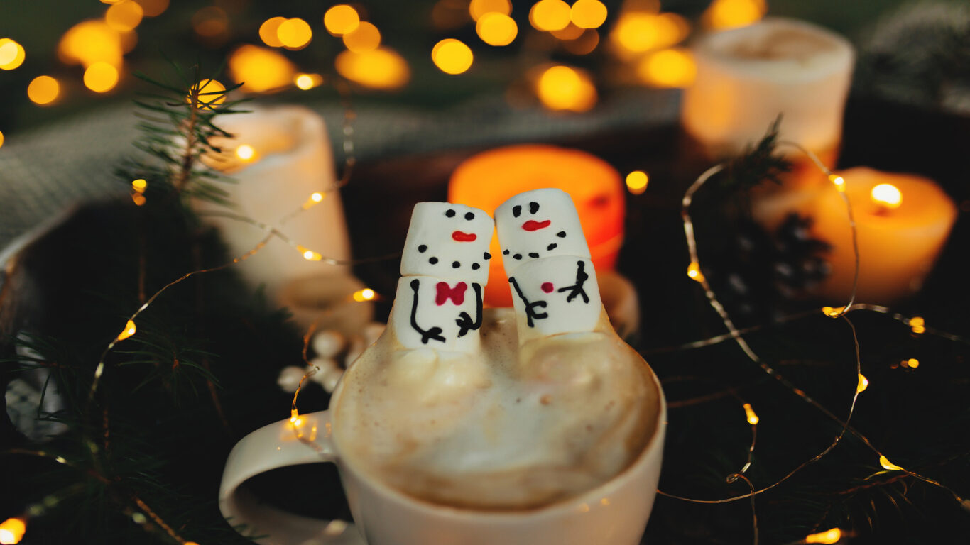 Two marshmallows decorated as snowmen sit in a mug of hot chocolate with whipped cream, surrounded by candles and string lights.