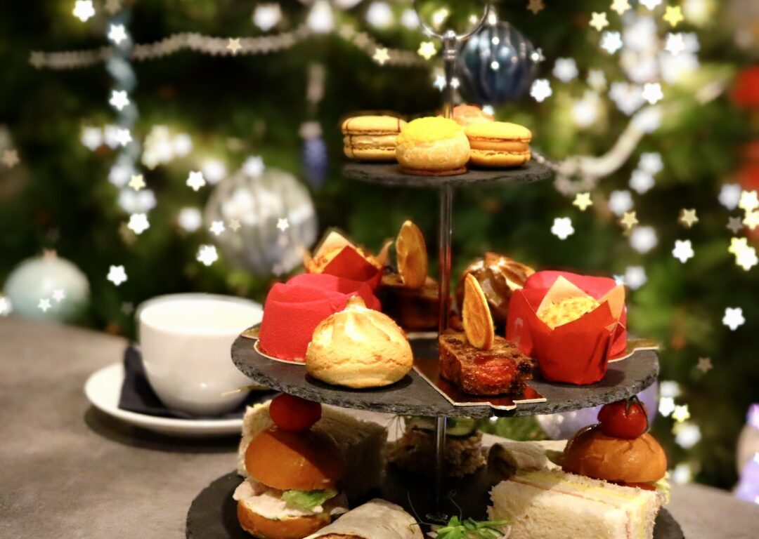 A three-tiered stand with assorted pastries, desserts, and finger sandwiches sits on a table in front of a decorated Christmas tree. A white teacup and saucer are beside the stand.