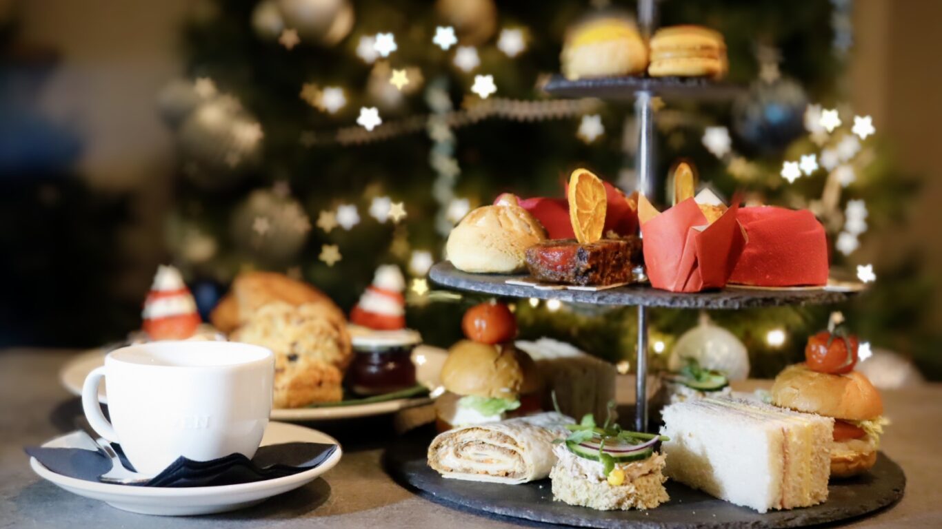 A three-tiered stand with assorted pastries, sandwiches, and desserts is set on a table next to a cup and saucer, with a decorated Christmas tree in the background.