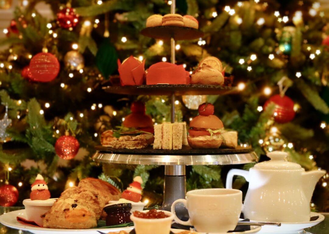 A tiered tray with assorted pastries, sandwiches, and desserts is set in front of a decorated Christmas tree, alongside cups, a teapot, and plates on a table.