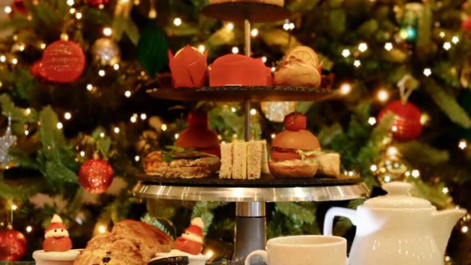 A tiered tray with assorted pastries, sandwiches, and desserts is set in front of a decorated Christmas tree, alongside cups, a teapot, and plates on a table.