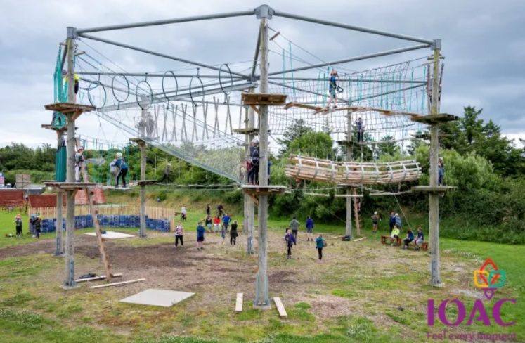 Children and adults participate in activities on an outdoor high ropes adventure course with nets, bridges, and platforms, surrounded by grass and trees.