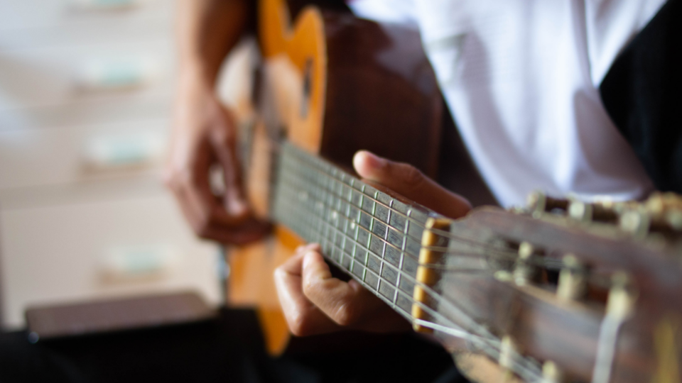 A person wearing a white shirt plays an acoustic guitar, focusing on the strings and fretboard with their left hand.